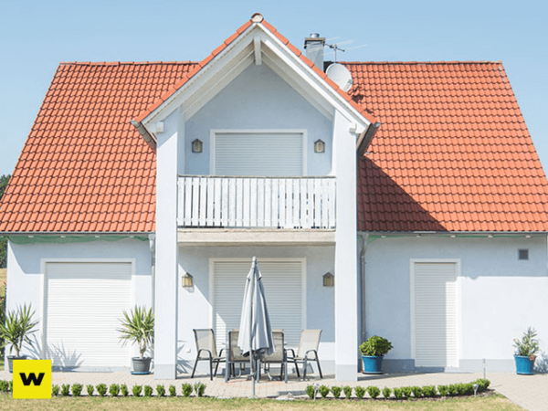 Modernes Einfamilienhaus mit rotem Dach, Terrasse und Rollläden bei sonnigem Wetter.